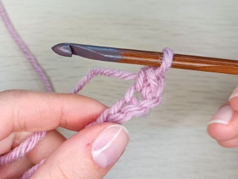 Close-up of hands performing a single crochet stitch with a wooden hook and purple yarn.