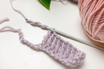 A small piece of purple crochet work with a crochet hook and strands of yarn on a white surface.