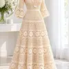 Ivory crochet lace maxi dress on dressmaker form with V neckline, bell sleeves, and three-tier flared skirt with floral rosette trim